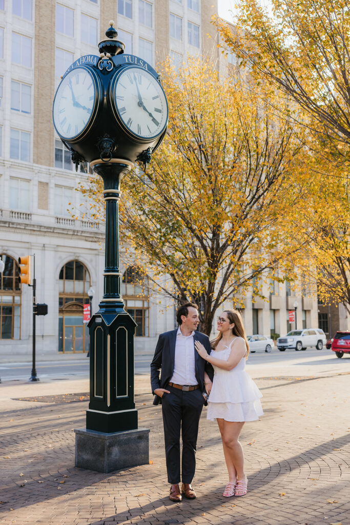 A couple stands in a tree-lined street; the man gently embraces the woman from behind. She smiles, looking at the camera in downtown tulsa for their engagement photos