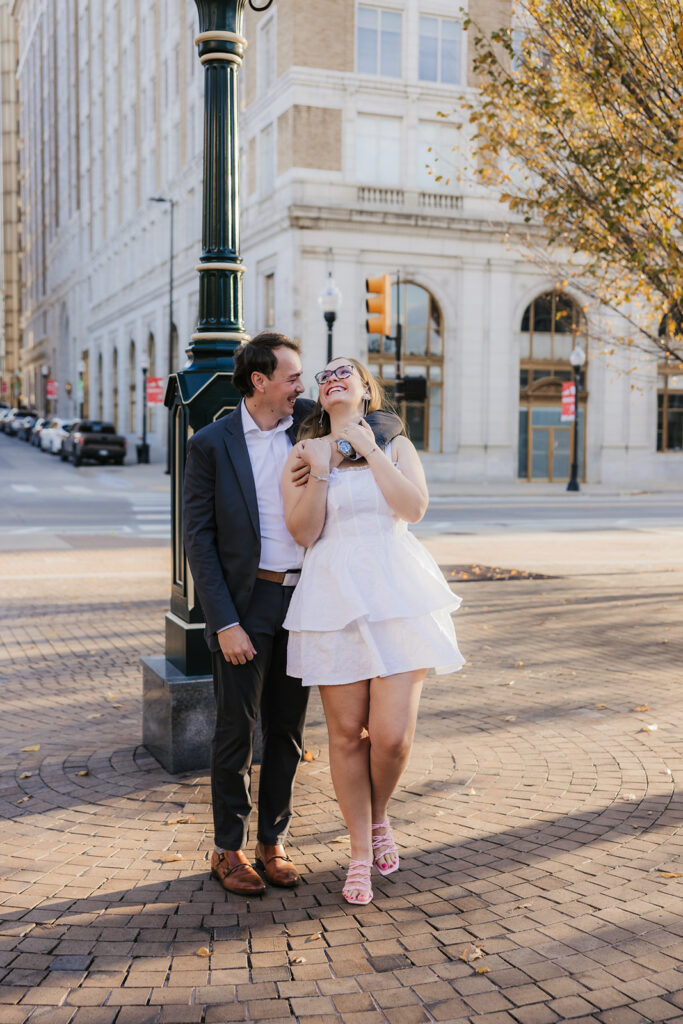 A couple stands in a tree-lined street; the man gently embraces the woman from behind. She smiles, looking at the camera in downtown tulsa for their engagement photos