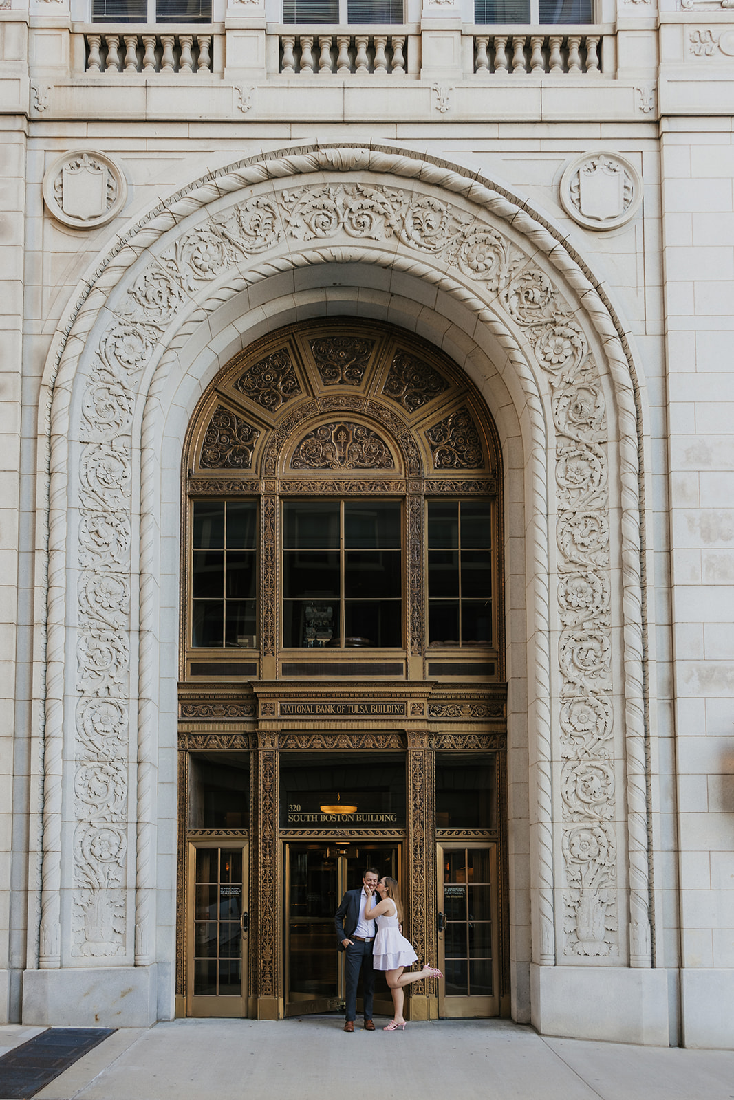 A couple stands together in front of a large ornate arched entrance of a stone building for their tulsa engagement photos