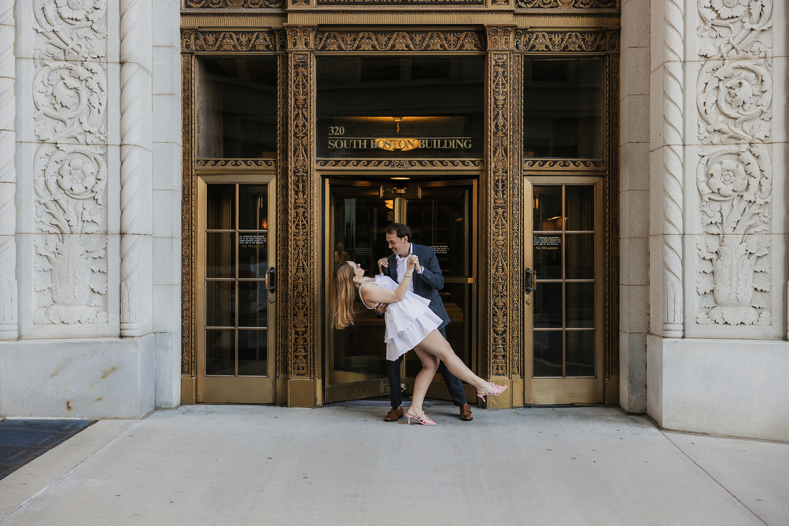 A couple stands together in front of a large ornate arched entrance of a stone building for their tulsa engagement photos