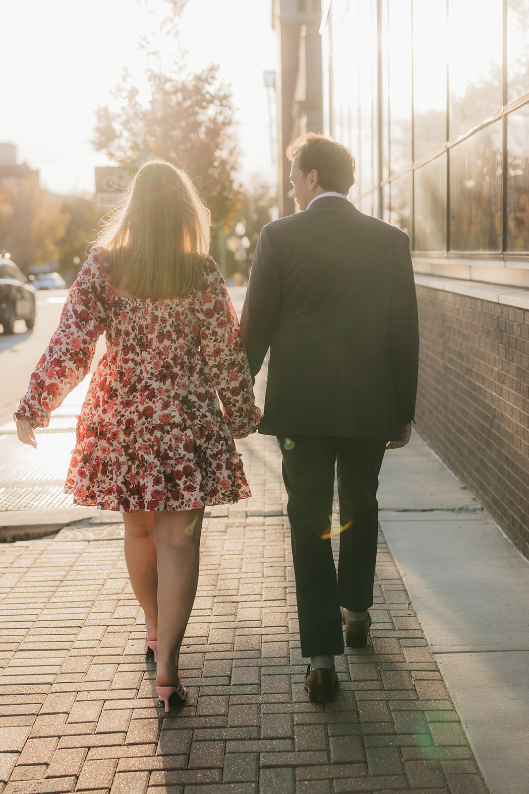 A couple walks hand in hand on a sunlit sidewalk, with their backs to the camera. The woman wears a floral dress, and the man is in a dark suit.