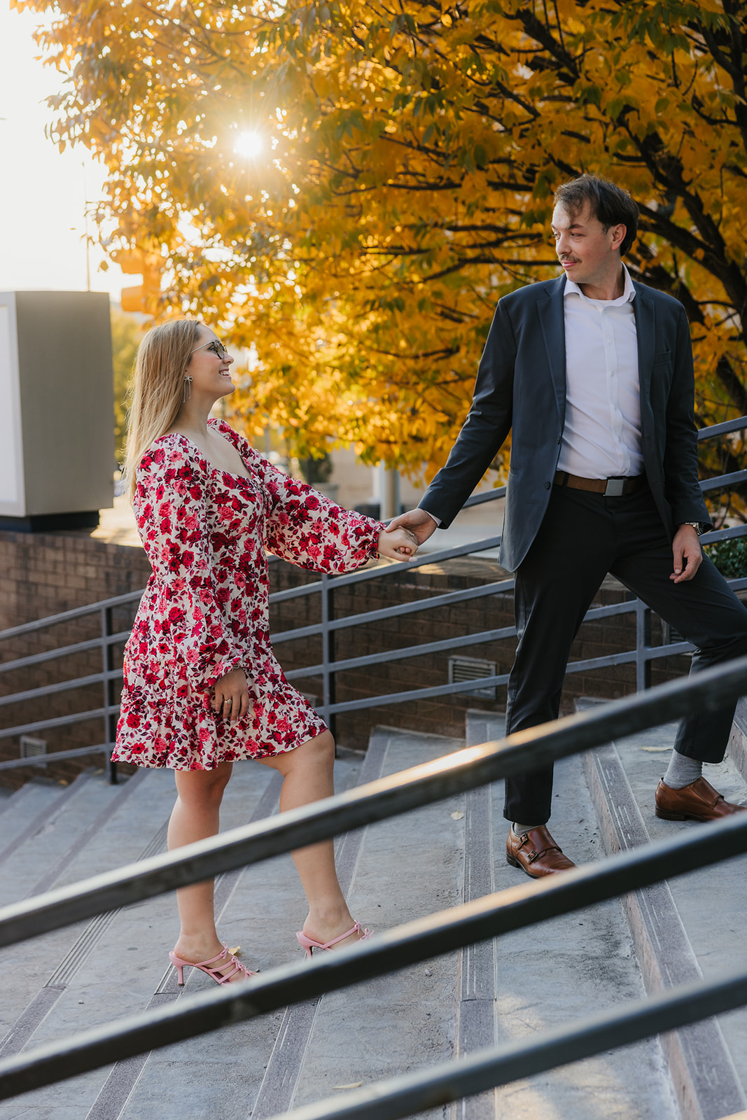 A couple walks hand in hand on a sunlit sidewalk, with their backs to the camera. The woman wears a floral dress, and the man is in a dark suit.