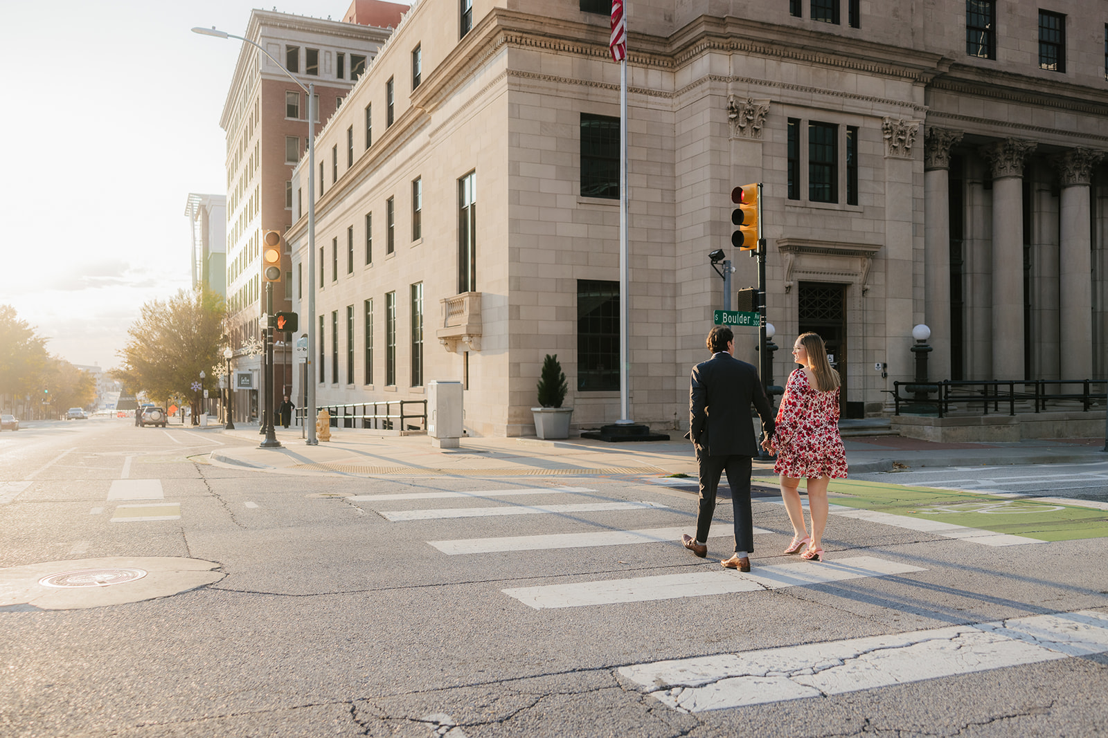 A couple walks hand in hand on a sunlit sidewalk, with their backs to the camera. The woman wears a floral dress, and the man is in a dark suit.