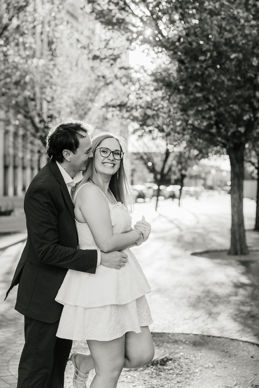 A couple stands in a tree-lined street; the man gently embraces the woman from behind. She smiles, looking at the camera in downtown tulsa for their engagement photos