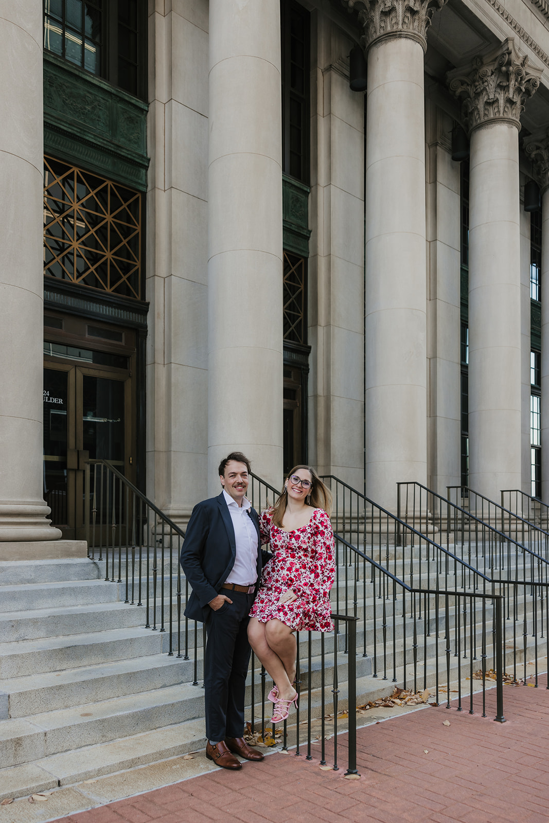 A couple walks hand in hand on a sunlit sidewalk, with their backs to the camera. The woman wears a floral dress, and the man is in a dark suit.