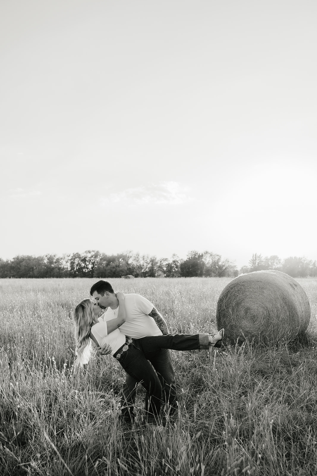 A man and woman stand in a grassy field near a large hay bale at sunset. Both are wearing white shirts and jeans.