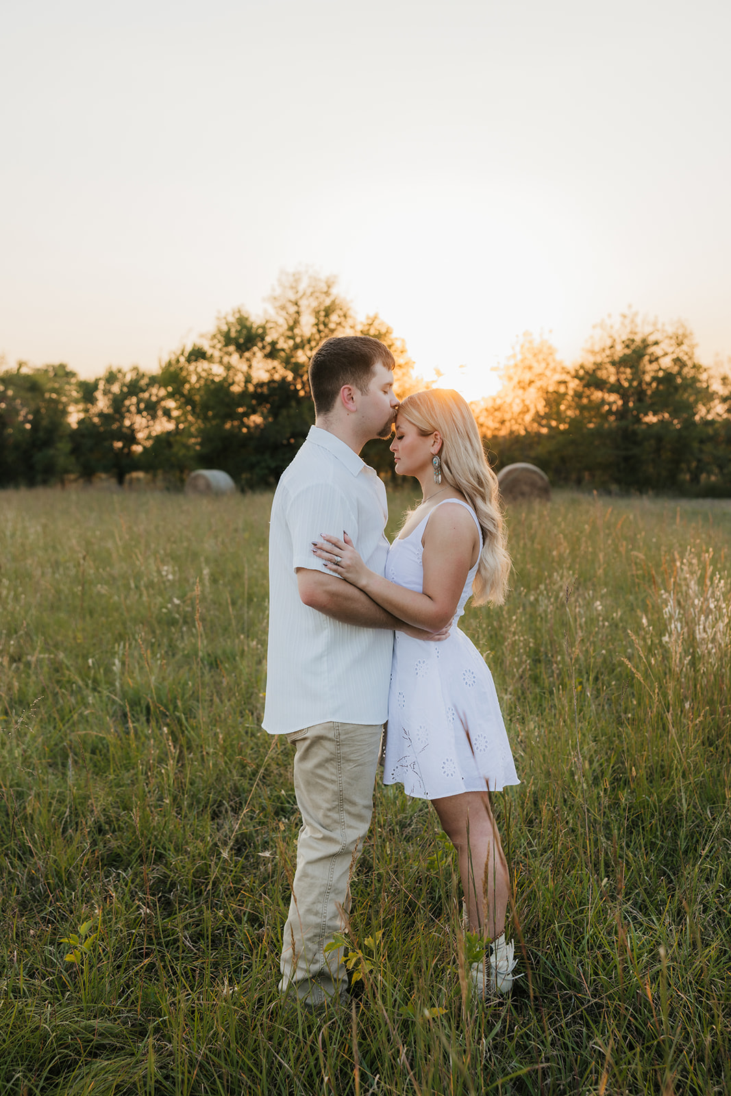 A couple embraces in a grassy field at sunset, with trees and a soft orange sky in the background.