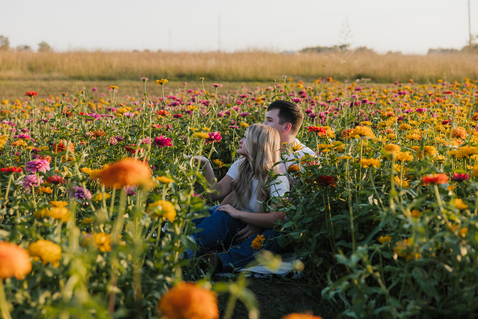 A couple sits in a field of vibrant flowers, surrounded by yellow and pink blooms, under a clear sky.