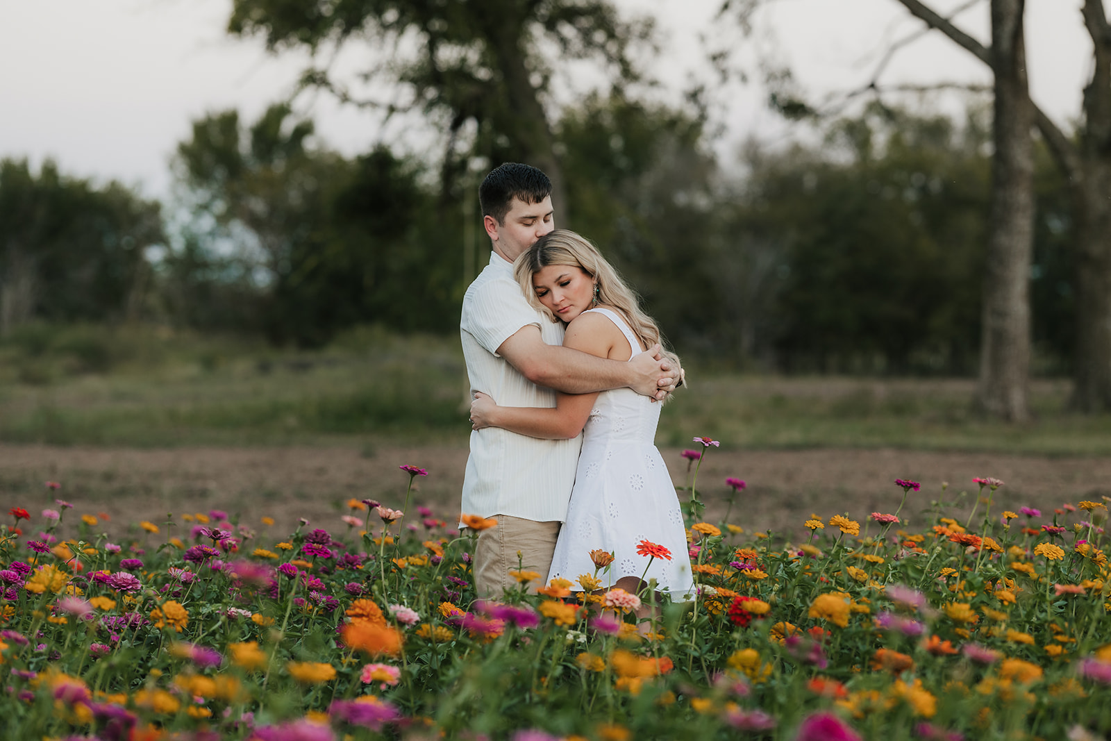 A couple holds hands while walking through a field of colorful flowers, with trees in the background under a clear sky.