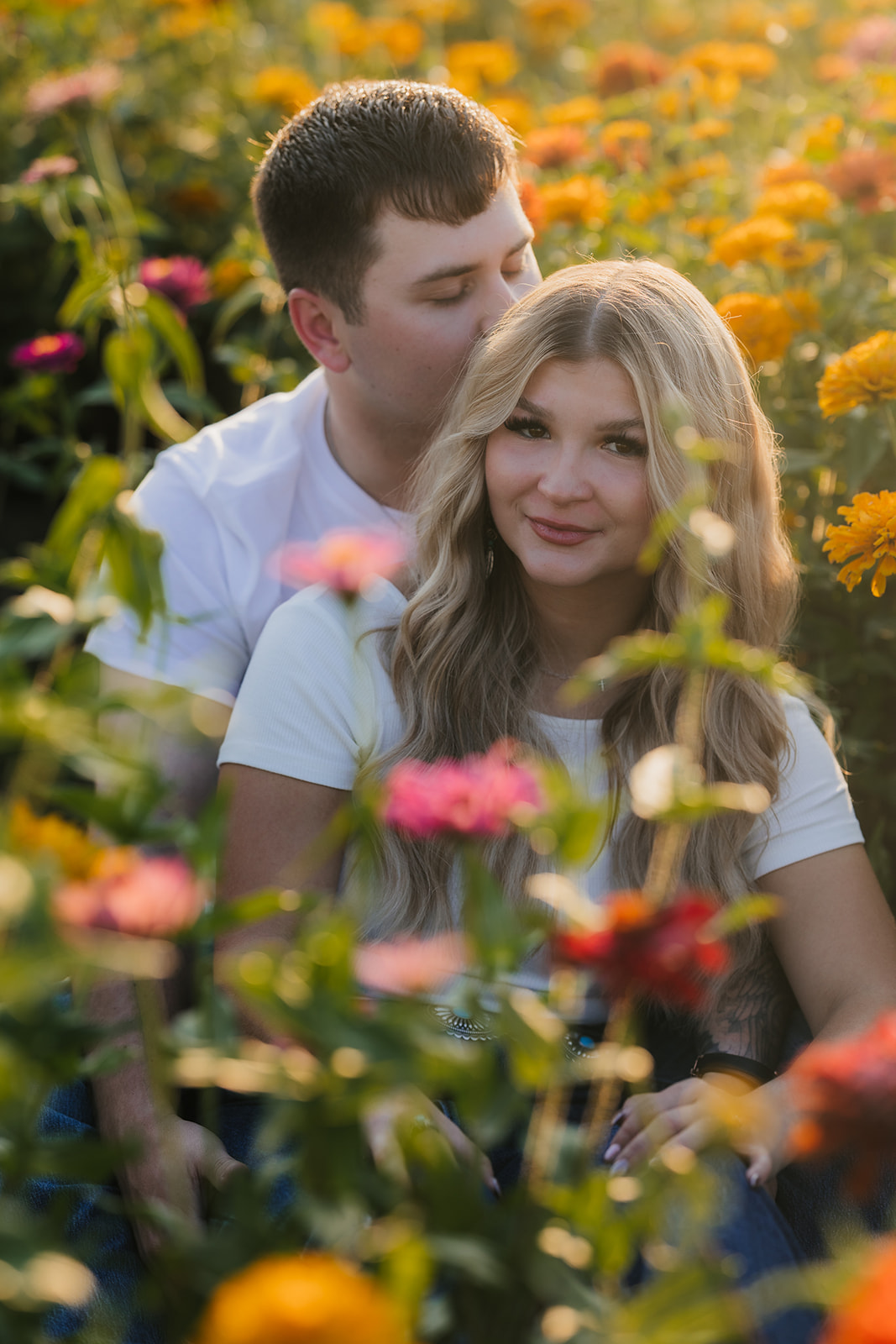 A couple sits together in a colorful flower field. The man is behind, leaning his face close to the woman's head. Both are wearing white shirts.