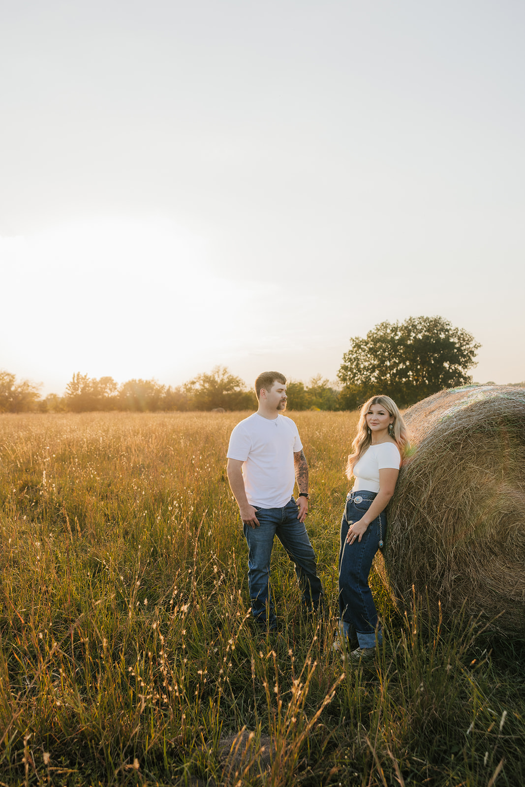 A man and woman stand in a grassy field near a large hay bale at sunset. Both are wearing white shirts and jeans.