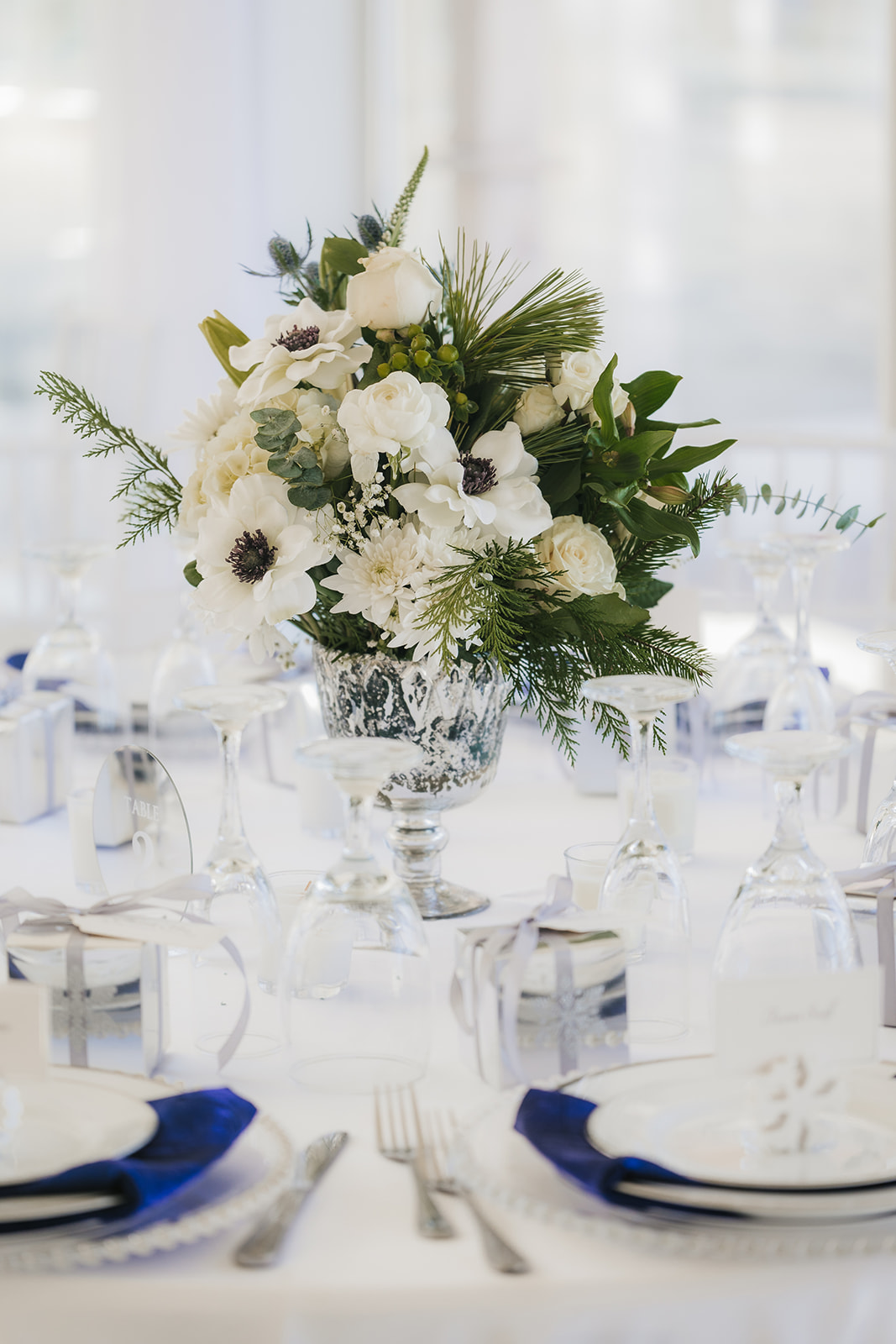 A floral centerpiece on a round table set with white and blue napkins, glassware, and silverware.