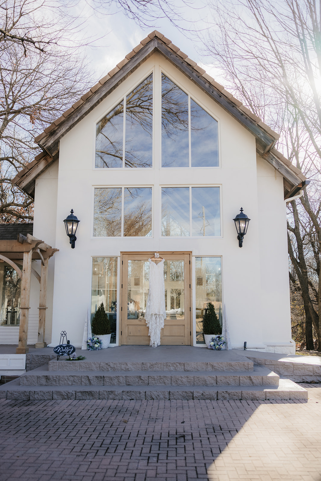 A white chapel with large glass windows, a wooden door, and lanterns on each side, set against a backdrop of leafless trees and a clear skyat glass chapel