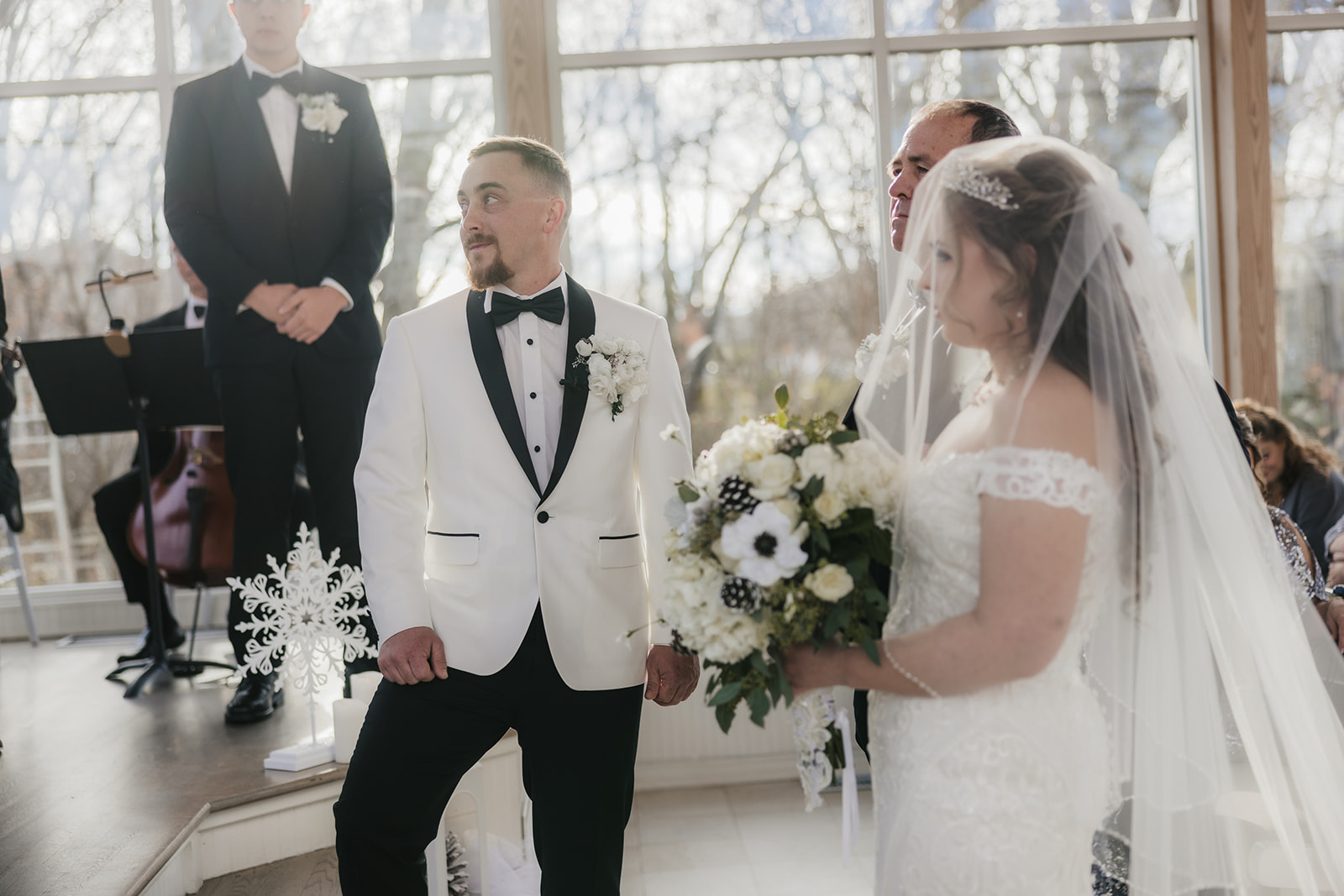 A bride and groom stand facing each other at an indoor wedding ceremony. The groom wears a white tuxedo, and the bride holds a bouquet of flowers. A man stands nearby, officiating at glass chapel