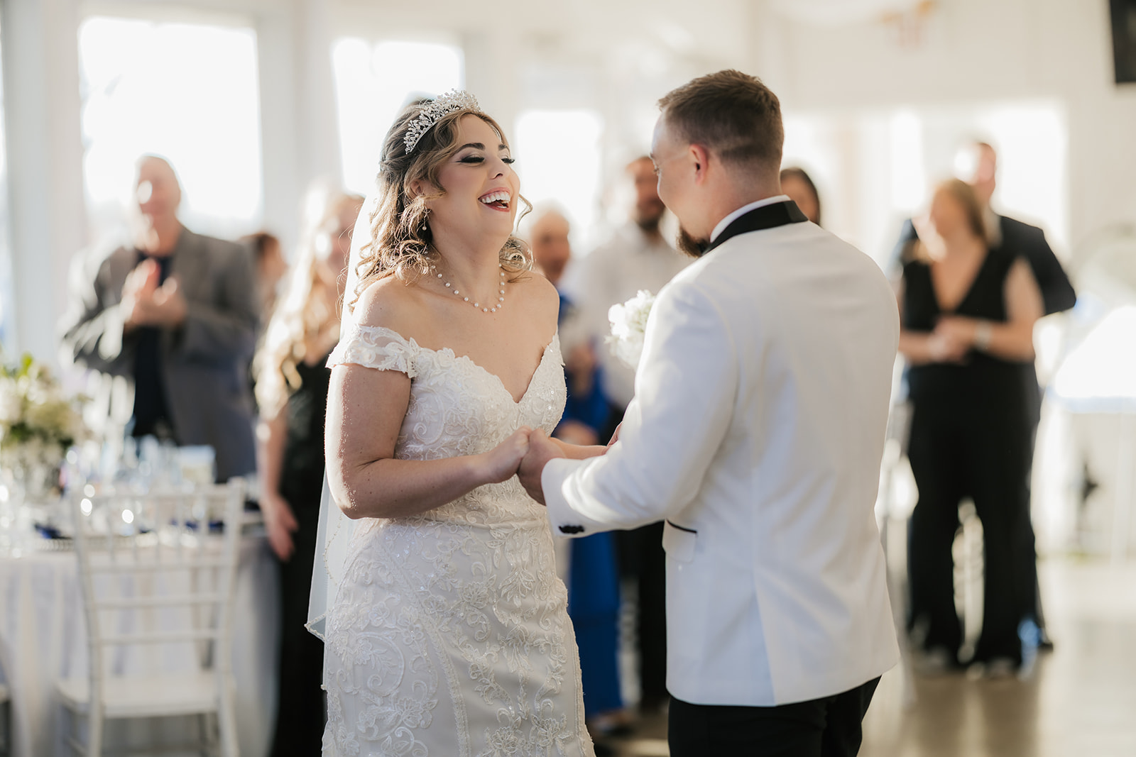 A bride and groom stand facing each other indoors. The bride is in a white dress, and the groom is in a white jacket. Guests are standing and sitting in the background at glass chapel