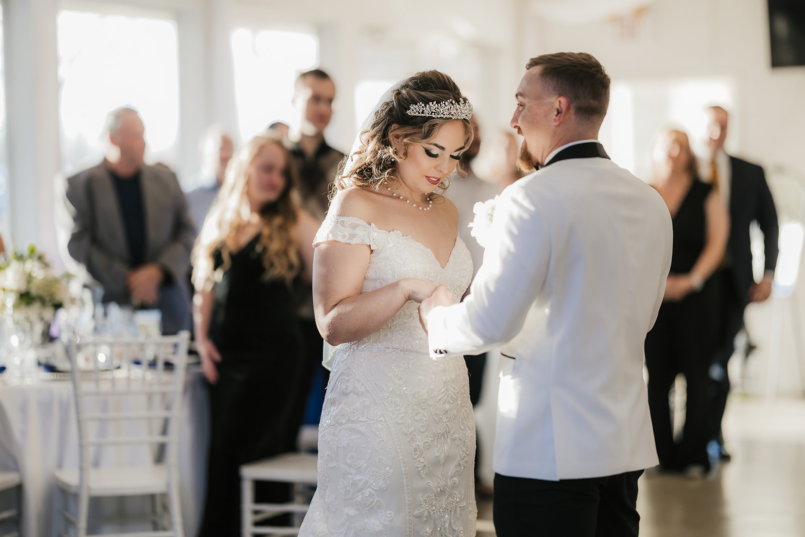 A bride and groom stand facing each other indoors. The bride is in a white dress, and the groom is in a white jacket. Guests are standing and sitting in the background at glass chapel