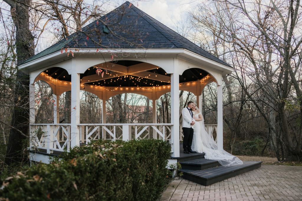 A couple in wedding attire stands on the steps of a white gazebo adorned with string lights, surrounded by trees without leaves.