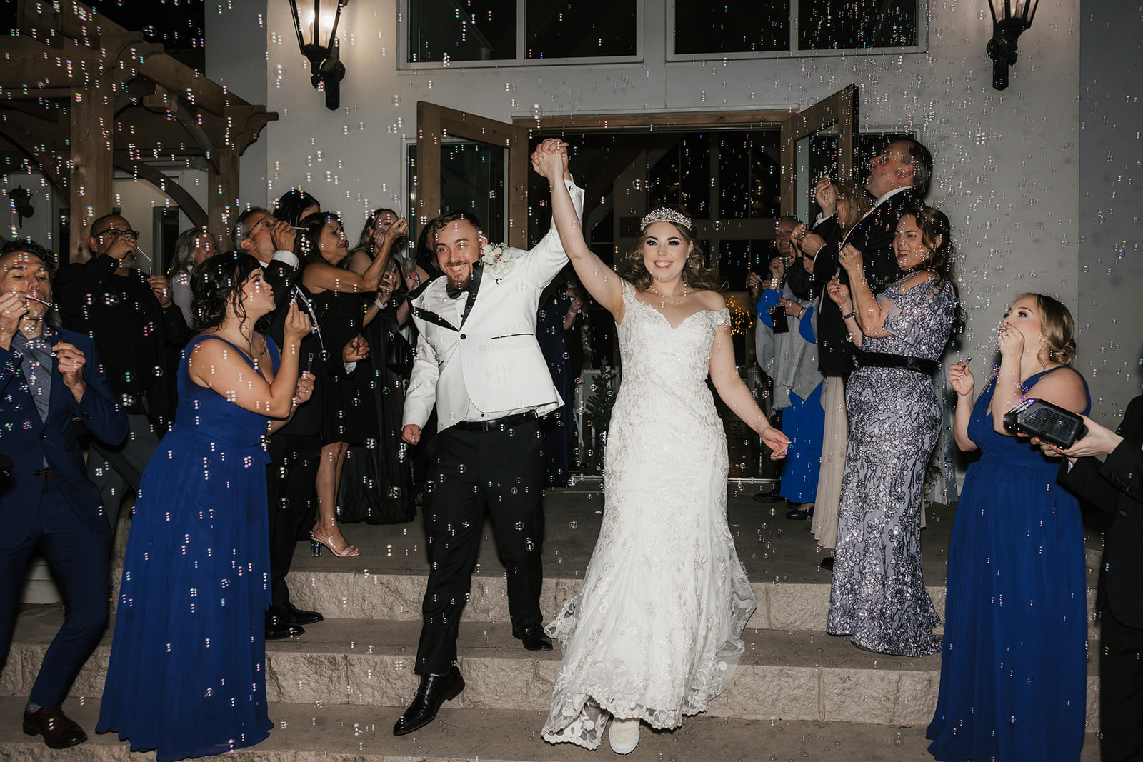 A bride and groom hold hands, smiling as they exit a building surrounded by guests blowing bubbles and clapping at glass chapel