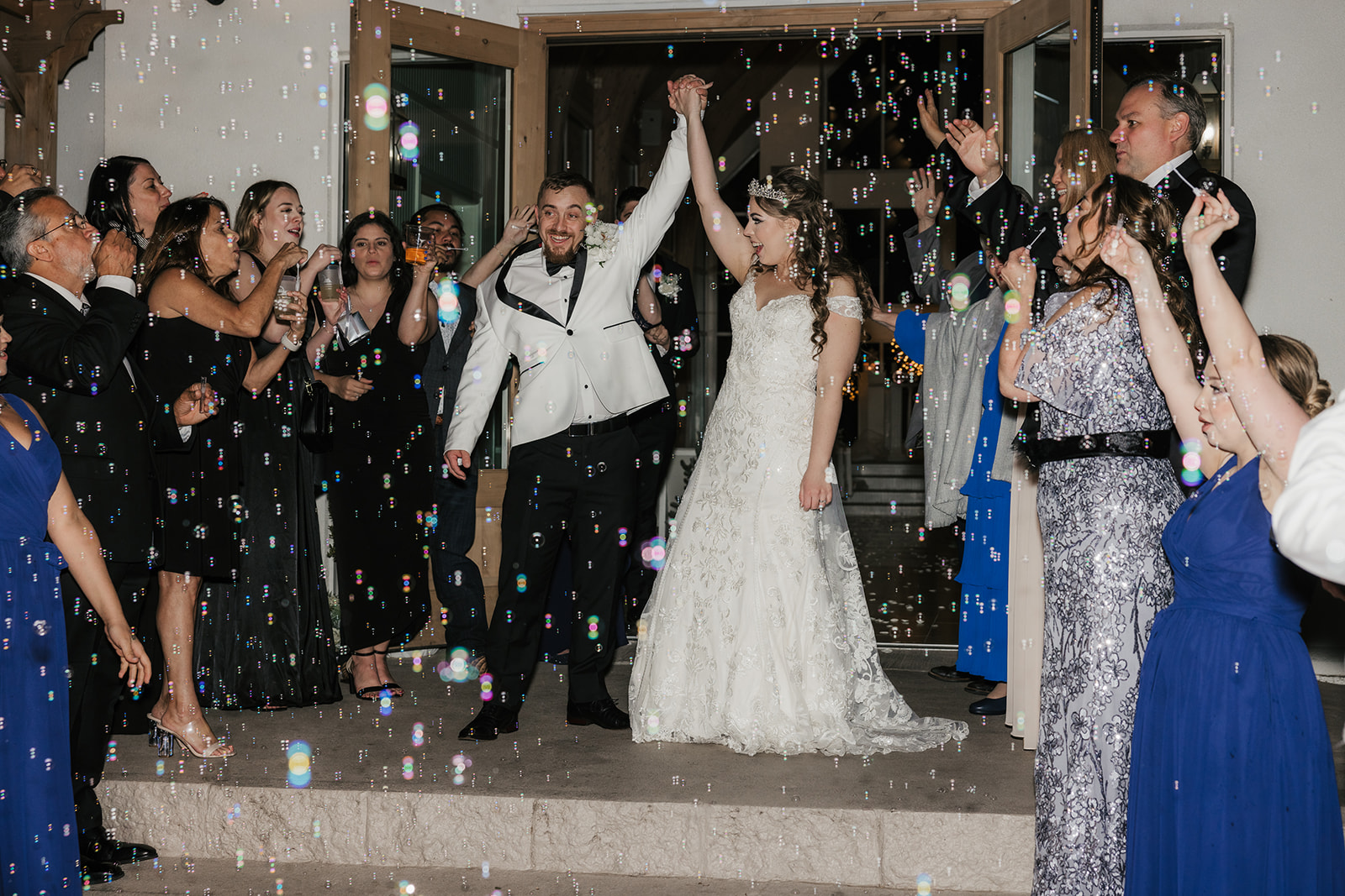 A bride and groom hold hands, smiling as they exit a building surrounded by guests blowing bubbles and clapping at glass chapel