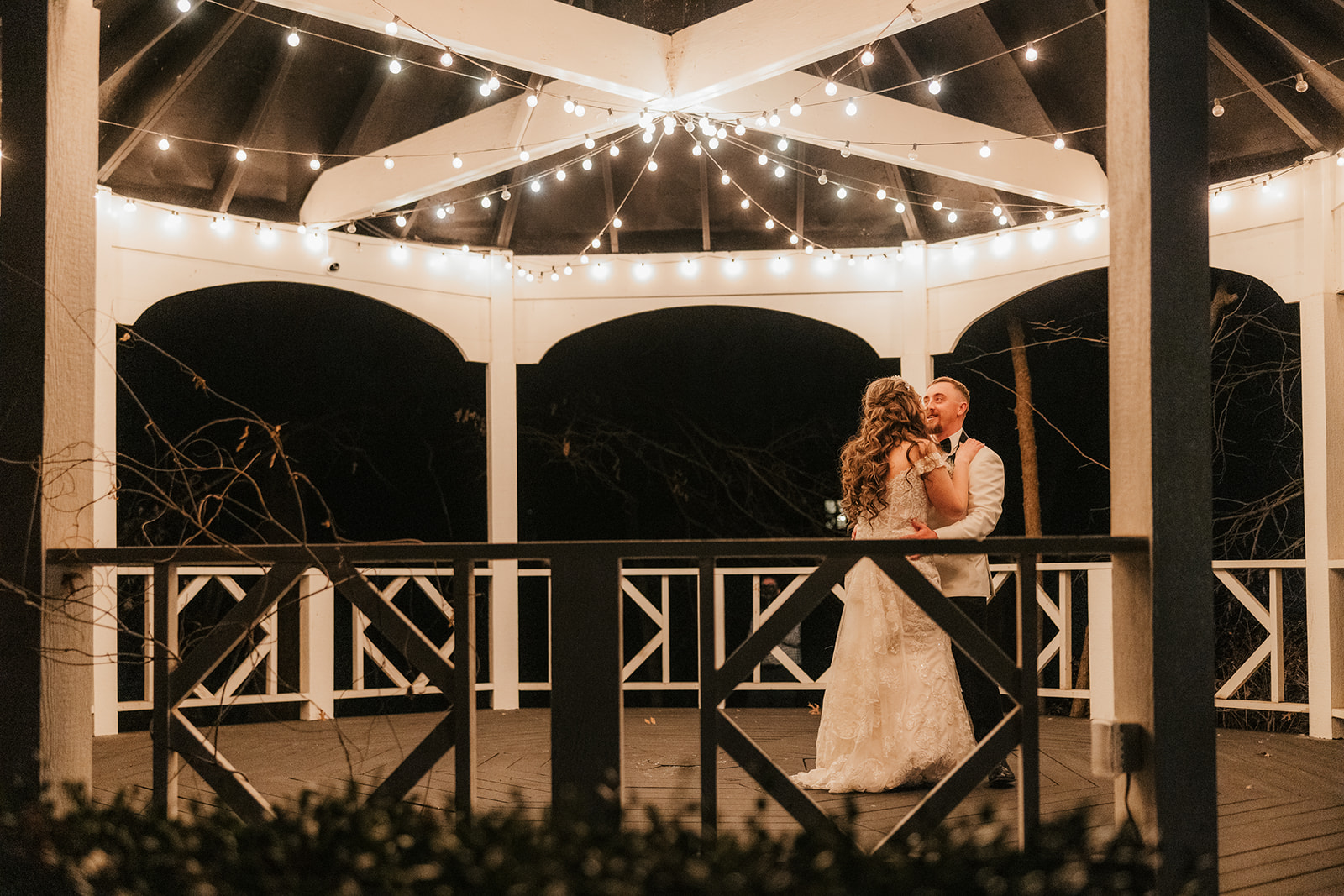 A couple dances under string lights in a gazebo at night. The bride wears a gown, and the groom is in a vest at glass chapel