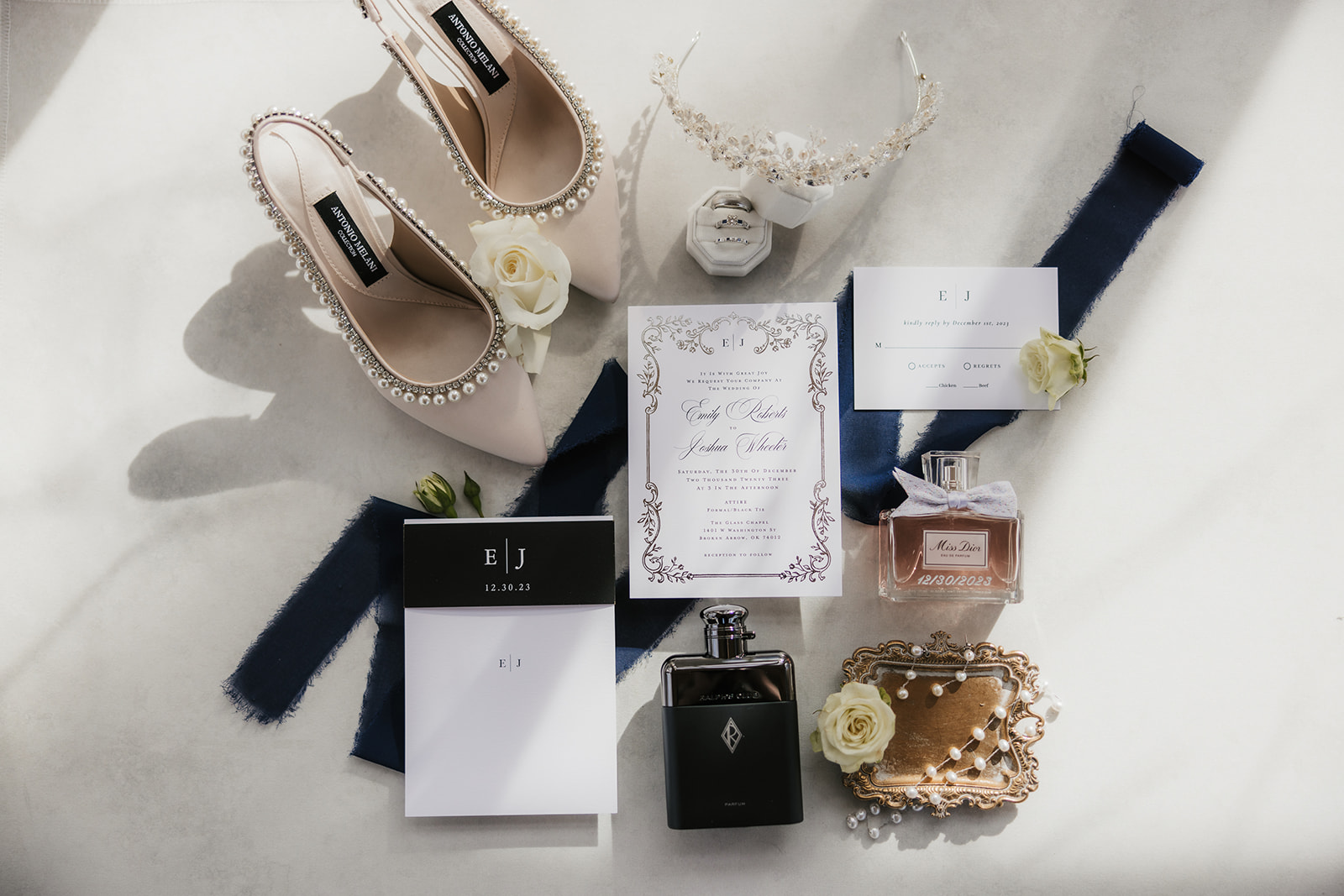 Flat lay of wedding accessories: shoes, perfume bottles, jewelry, invitation, and cards on a white background decorated with roses.