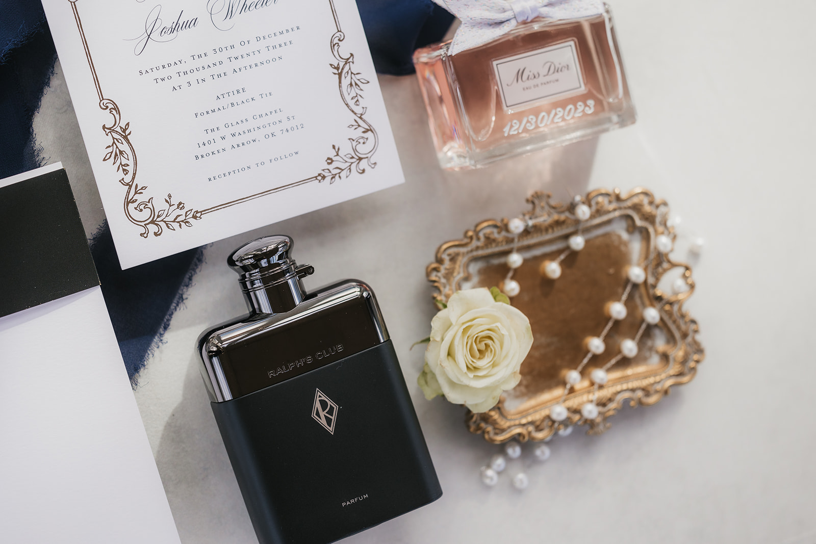 Wedding invitation set with ornate design, perfume bottle, and roses arranged on a white surface with a blue ribbon.