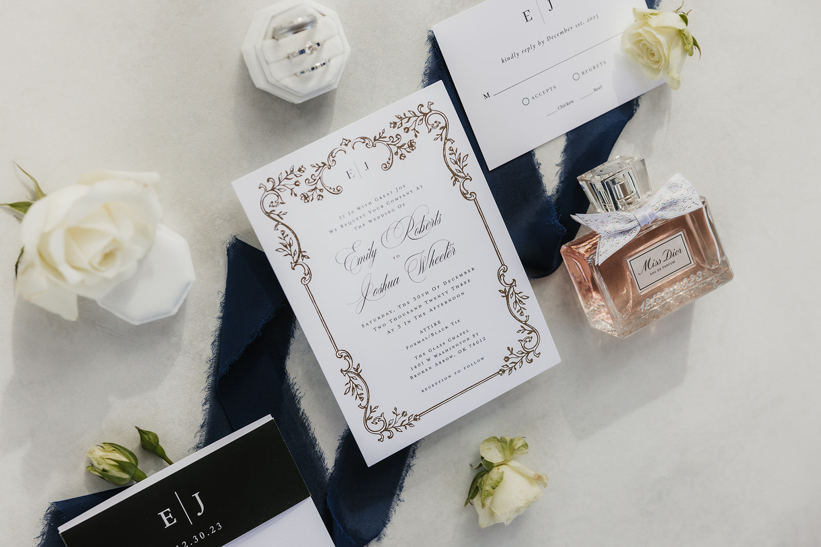 Wedding invitation set with ornate design, perfume bottle, and roses arranged on a white surface with a blue ribbon.