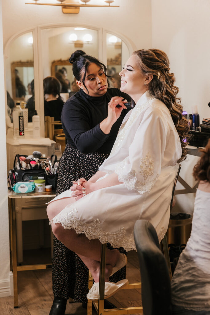 A makeup artist applies cosmetics to a woman seated in a robe in front of a vanity mirror.