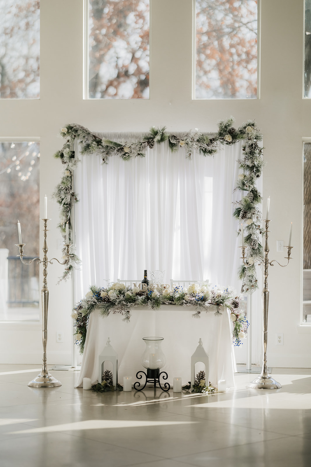 A decorated wedding altar with white drapery, floral garlands, and tall candelabras in a bright, sunlit room.