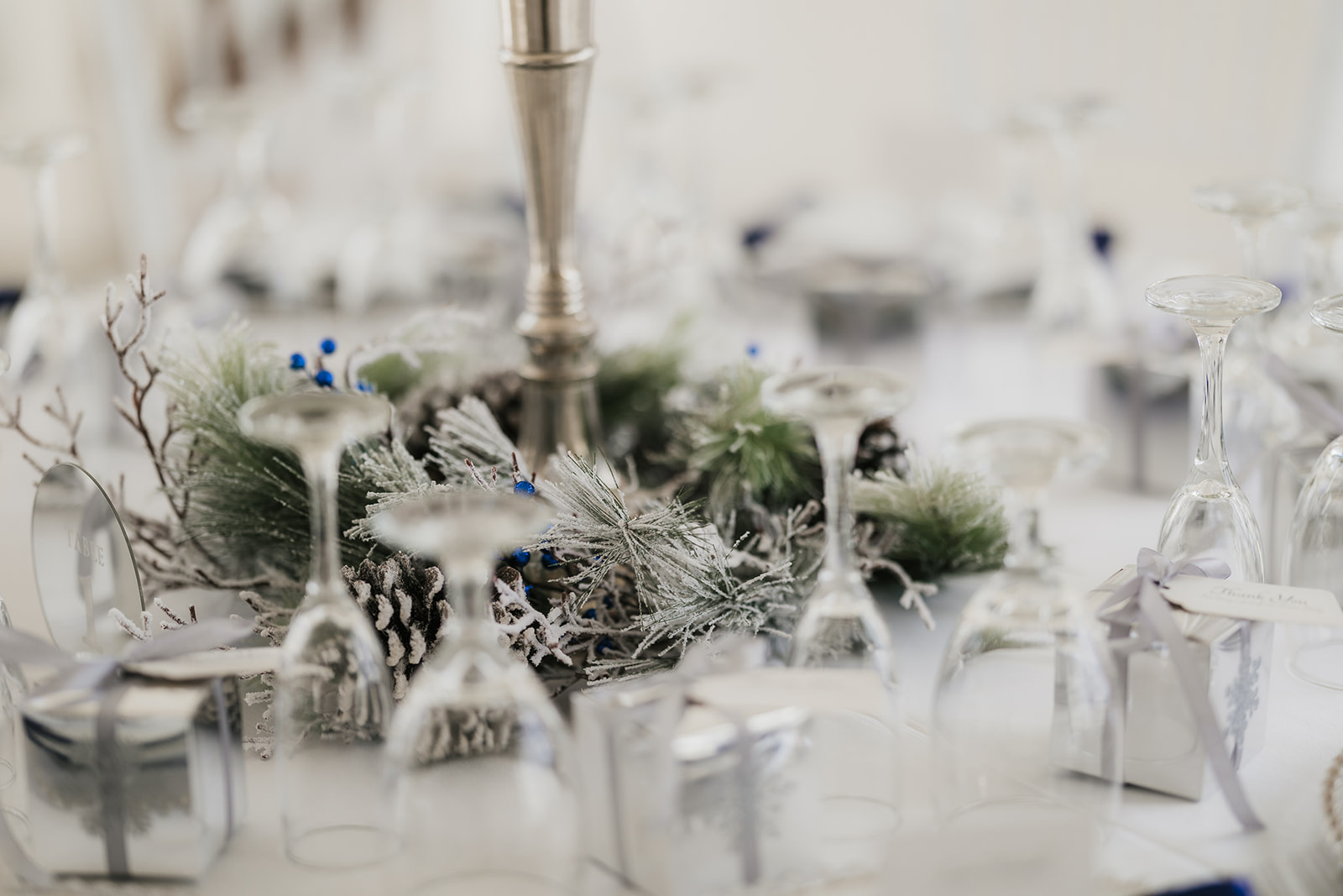 Elegant table setting with upside-down wine glasses, metallic gift boxes, and a centerpiece of pine branches and blue berries.