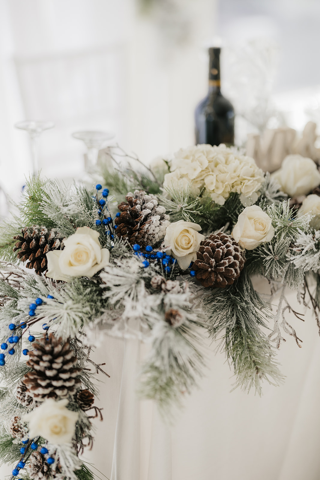 Winter-themed floral arrangement with white roses, pinecones, green branches, and blue berries. A wine bottle is in the background.