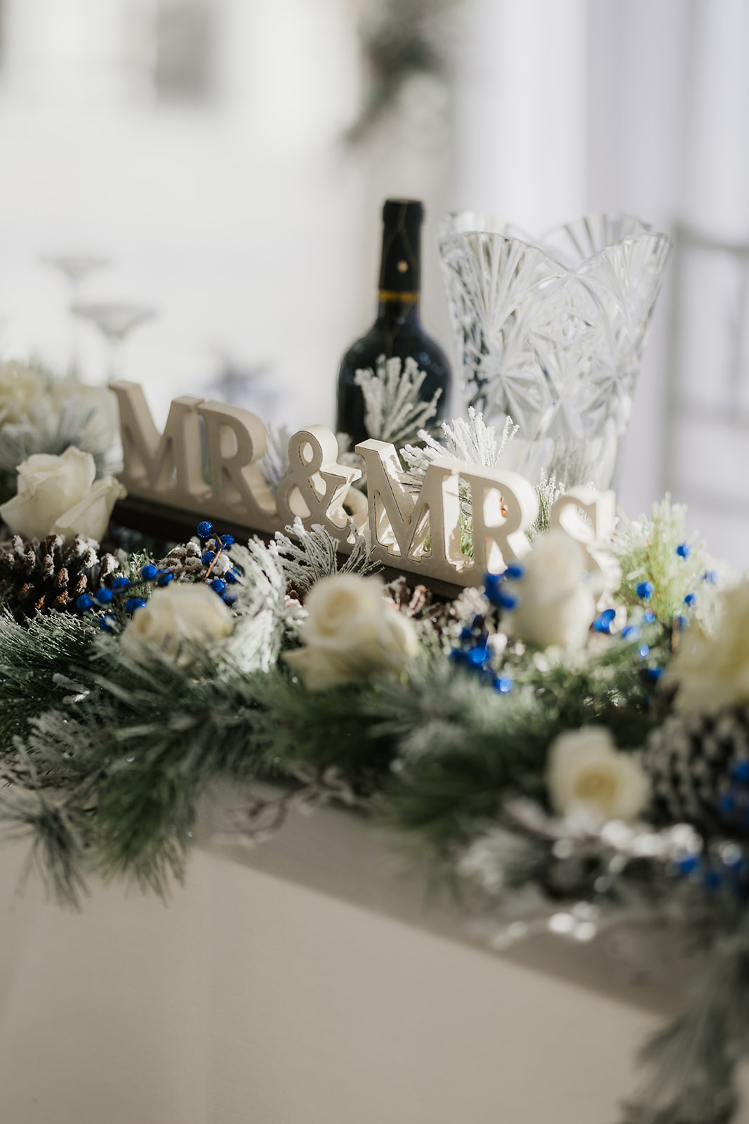 A winter-themed wedding table centerpiece with a "MR & MRS" sign, white roses, blue accents, pine branches, a wine bottle, and crystal glassware.