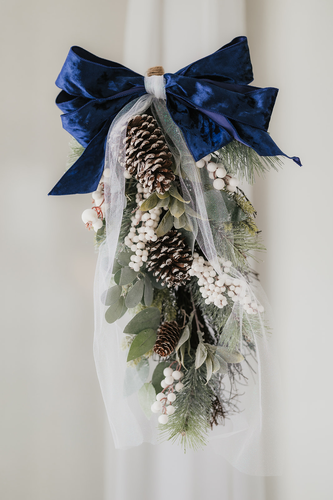 Decorative swag with pinecones, white berries, greenery, sheer white ribbon, and a large blue bow.