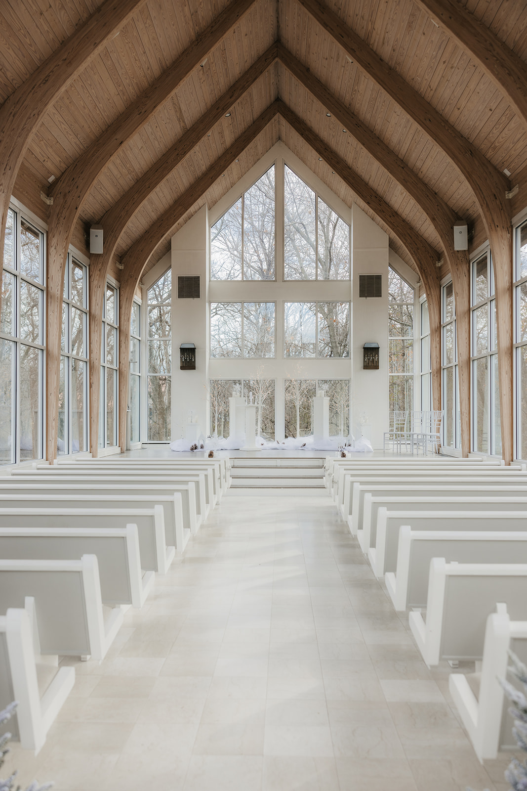 Interior of a bright, modern chapel with large windows, white pews, and small decorated trees lining the aisle at the glass chapel