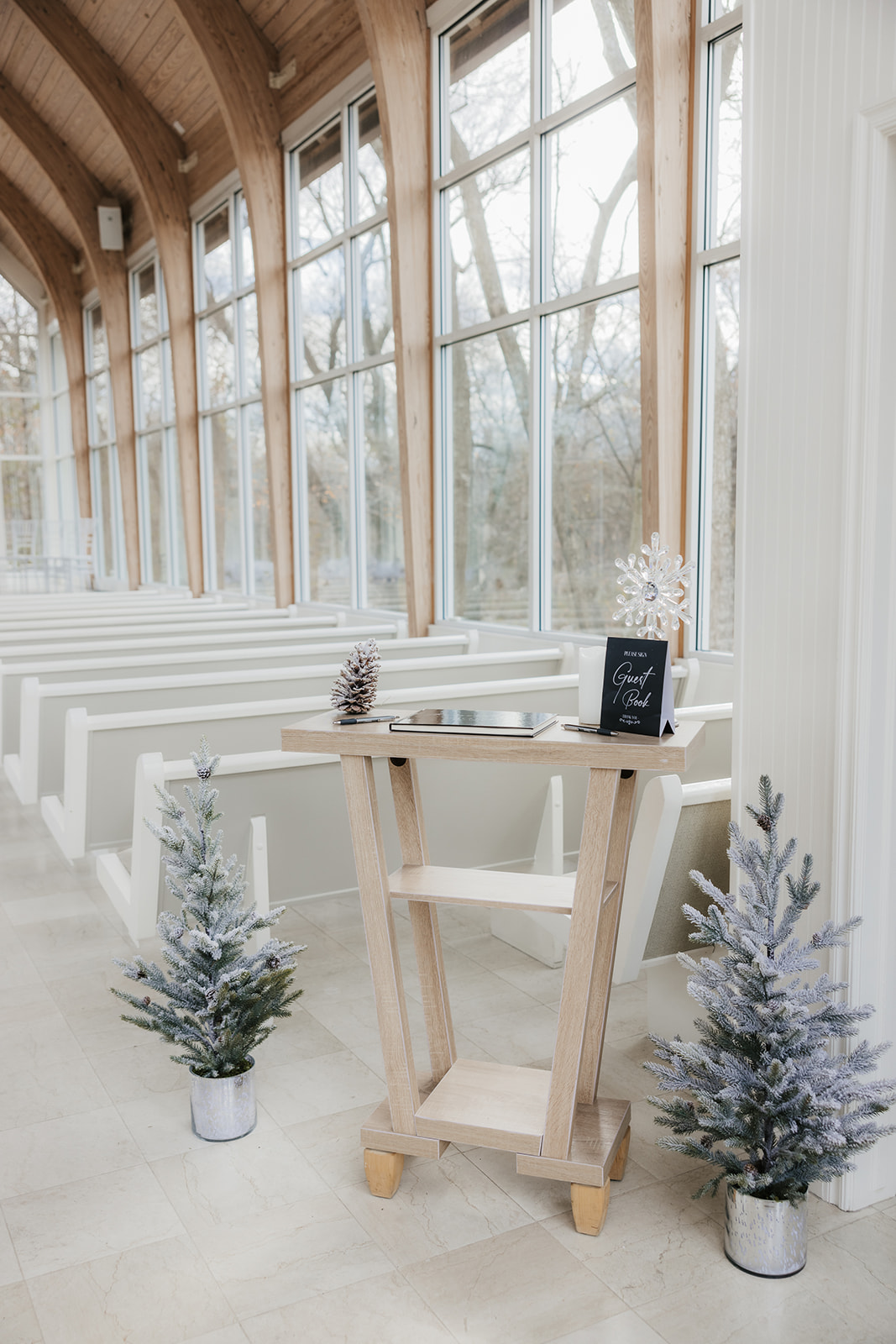 Interior of a bright, modern chapel with large windows, white pews, and small decorated trees lining the aisle at the glass chapel