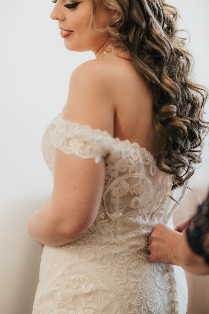 A bride in a lace wedding dress has her gown adjusted by another person.