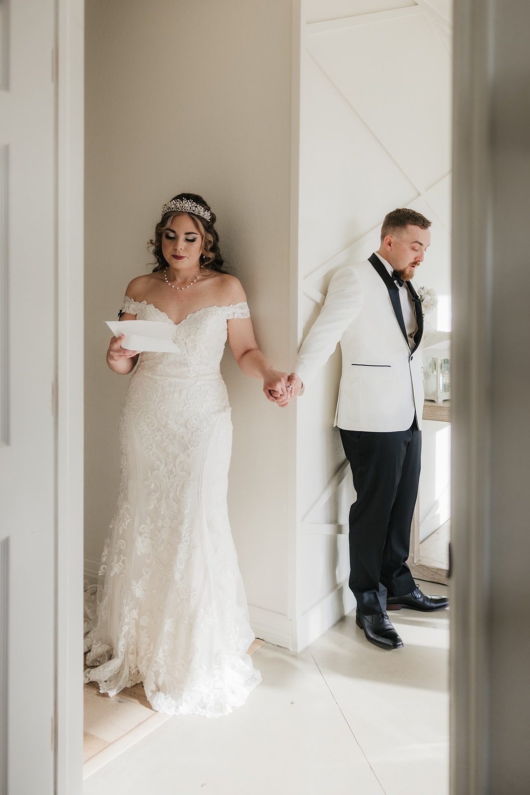 A bride and groom stand back to back, holding hands around a corner. The bride in a white dress reads a note while the groom in a white tuxedo jacket appears contemplative.