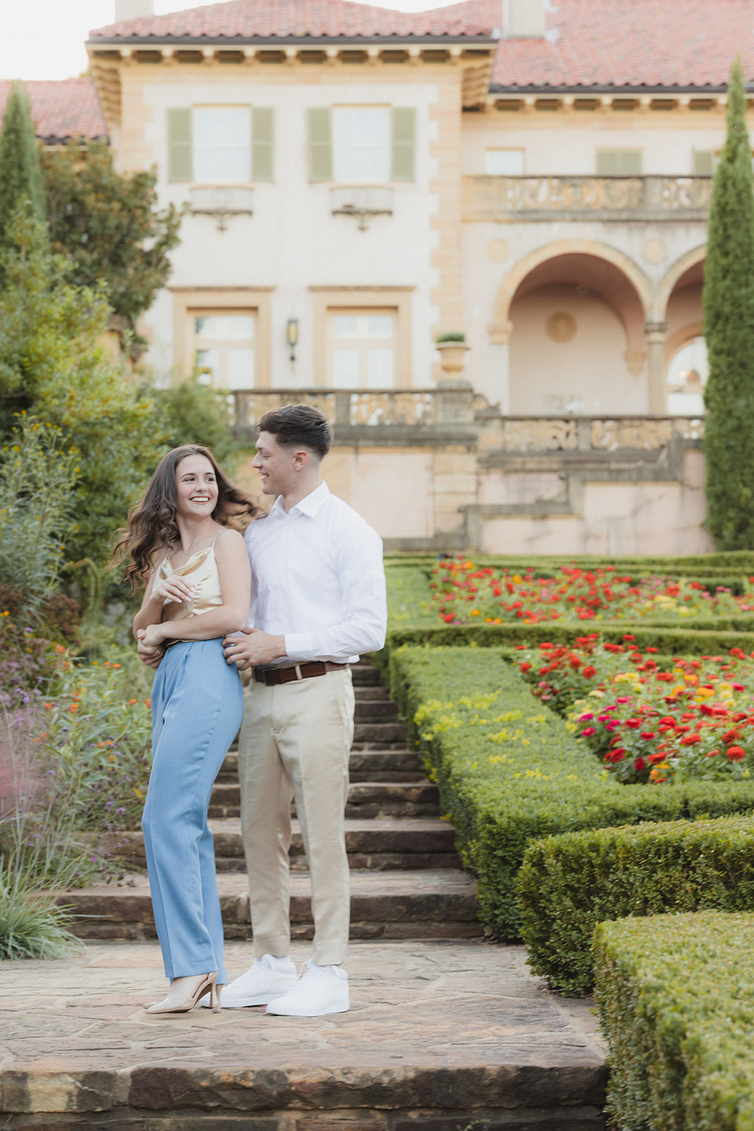 A couple stands and smiles on a stone path in a garden with vibrant flowers, in front of a large, ornate building for their tulsa engagement photos