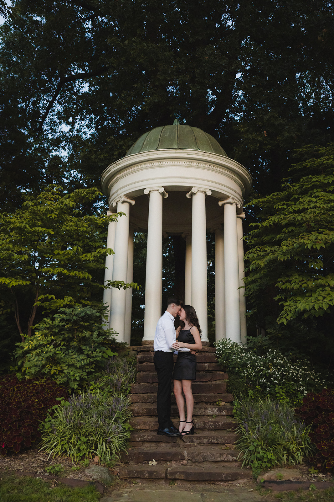 A couple embraces on stone steps in front of a small white columned gazebo surrounded by trees and greenery.