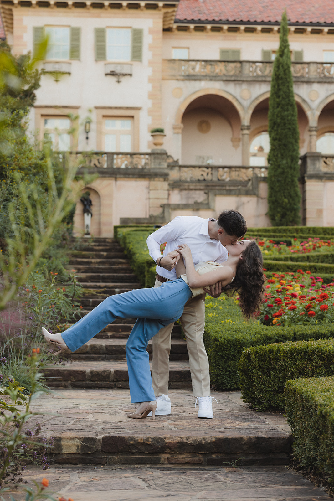 A couple stands and smiles on a stone path in a garden with vibrant flowers, in front of a large, ornate building for their tulsa engagement photos