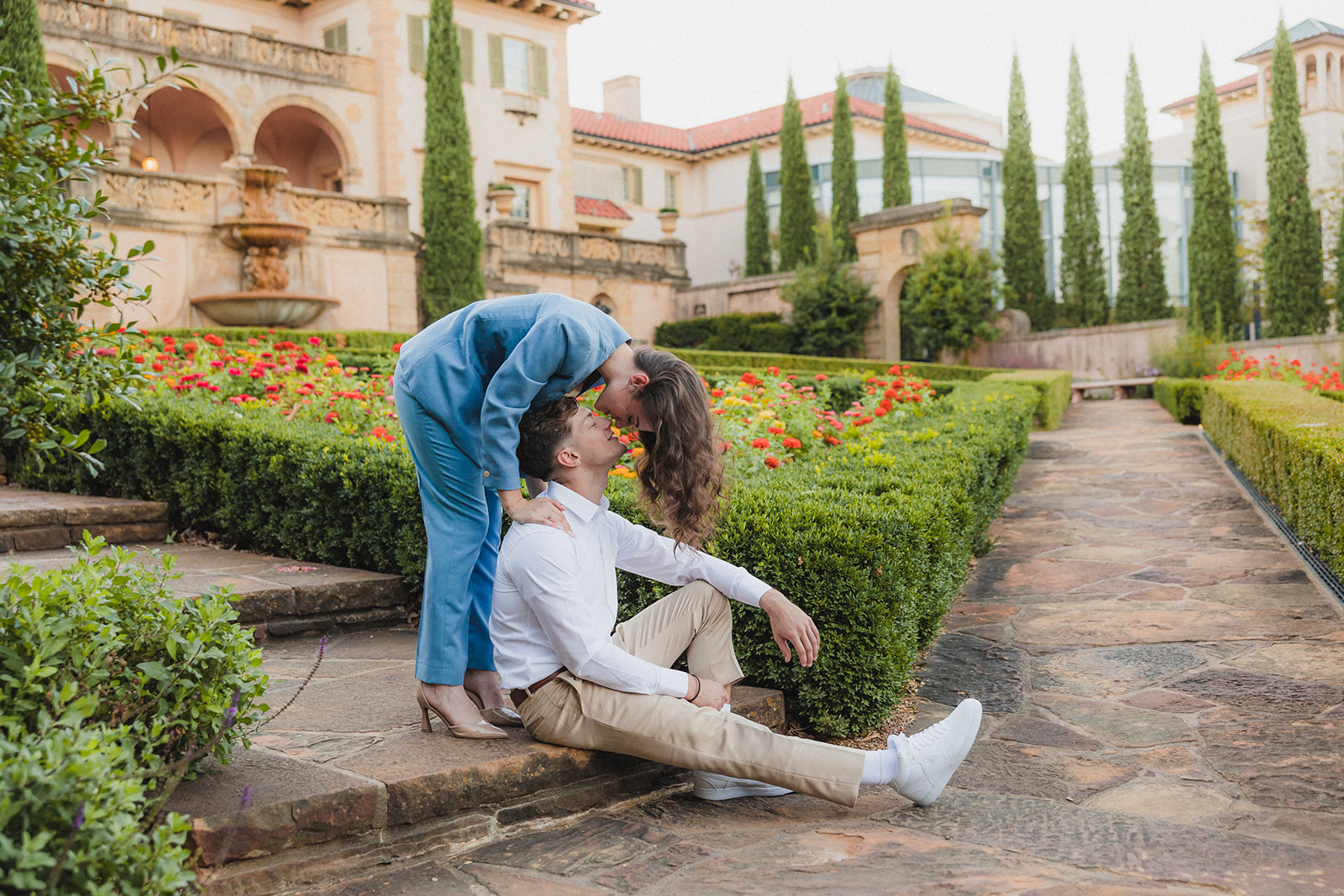 A couple stands and smiles on a stone path in a garden with vibrant flowers, in front of a large, ornate building for their tulsa engagement photos