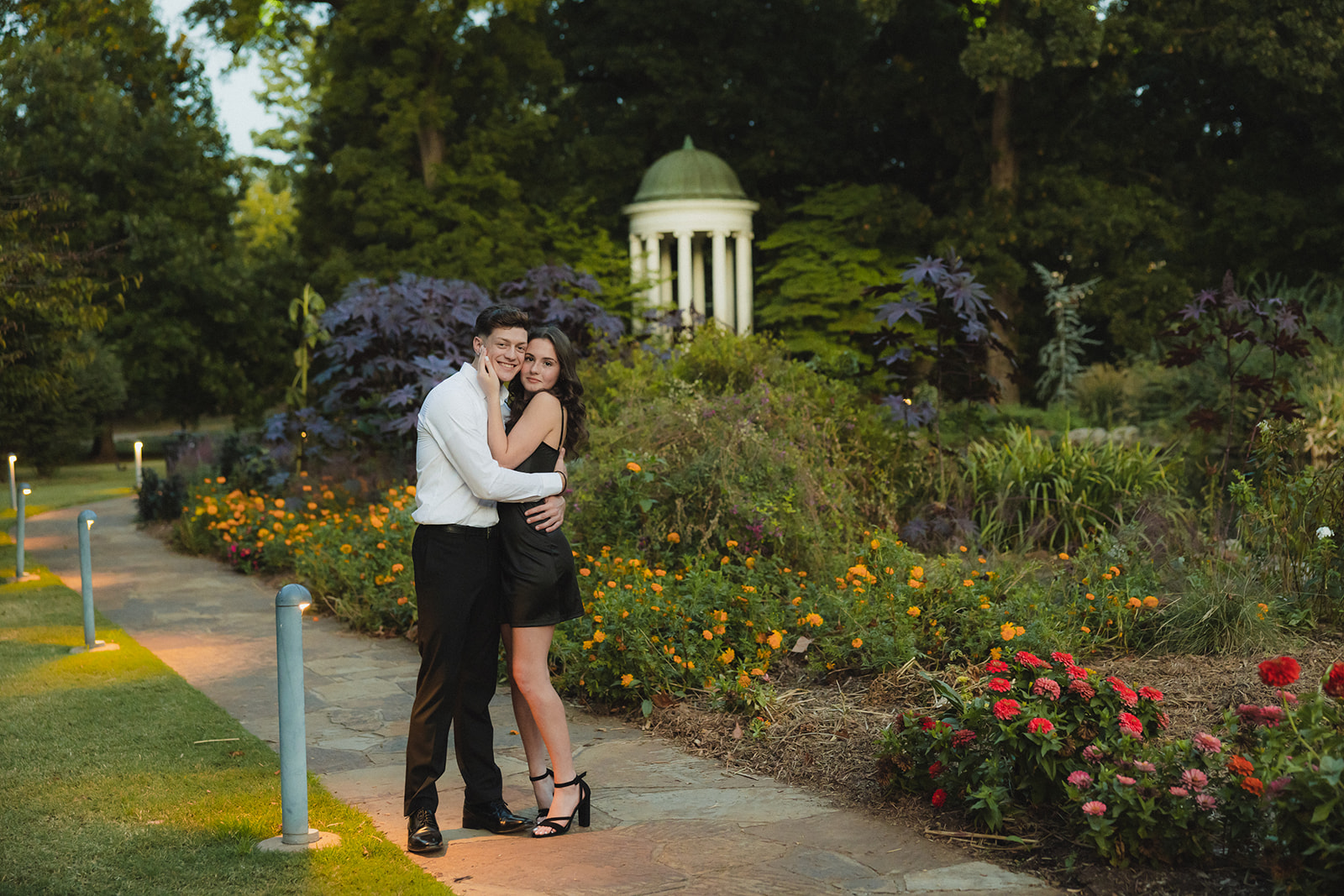 A couple embraces on a garden path, surrounded by flowers and greenery, with a small white gazebo in the background.