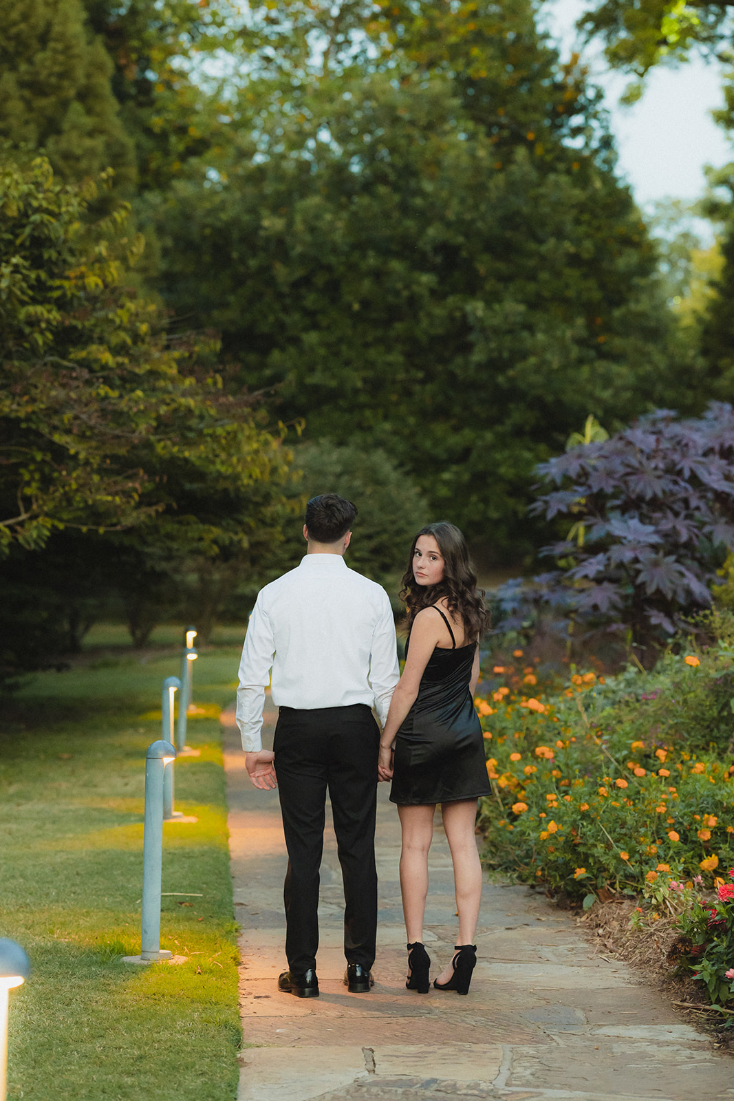 A couple embraces on a garden path, surrounded by flowers and greenery, with a small white gazebo in the background.