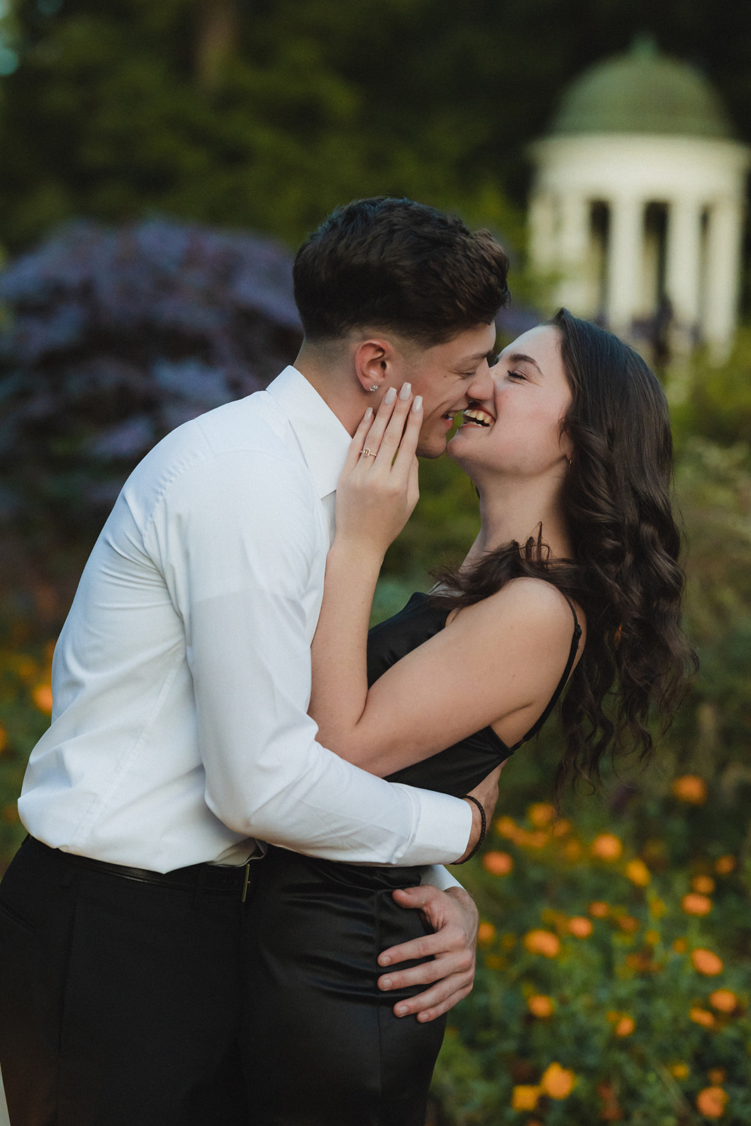 A couple stands close in a garden, smiling at each other, with a gazebo visible in the background.