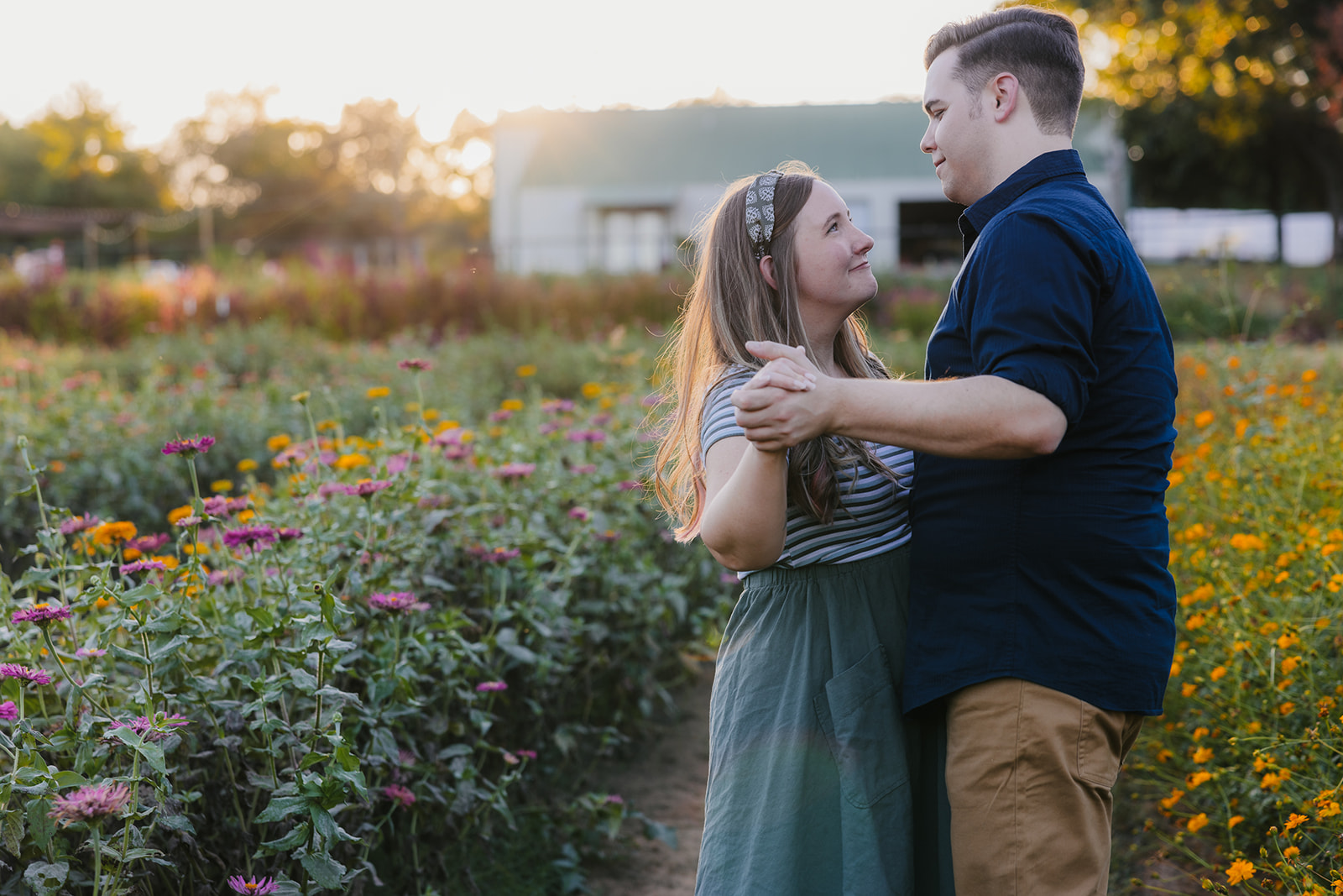 A couple embraces in a field of wildflowers during sunset, with trees and a structure visible in the background.