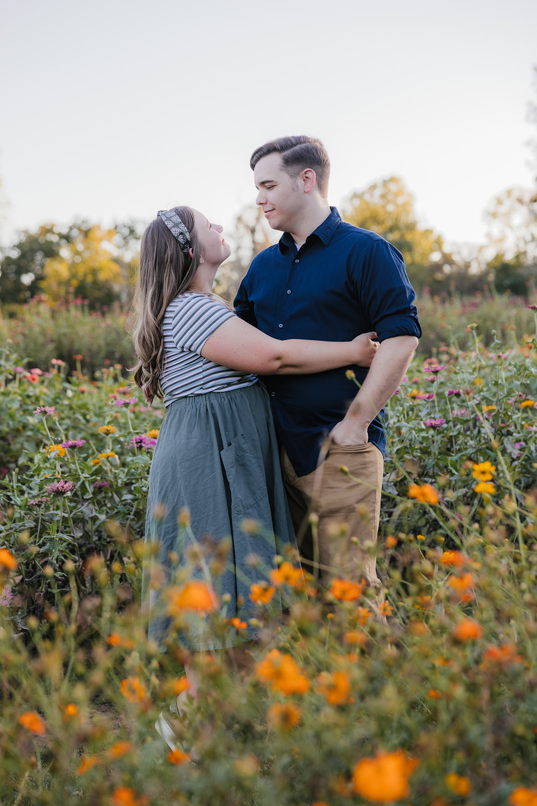 A couple embraces in a field of wildflowers during sunset, with trees and a structure visible in the background.