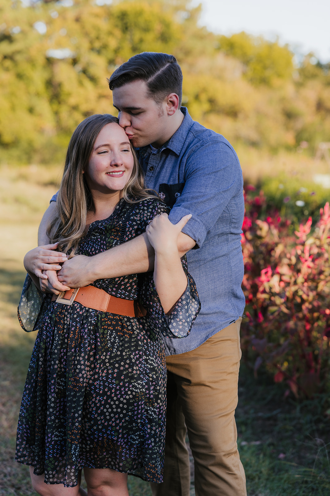 A man and woman stand in a garden, with the man hugging the woman from behind. The woman smiles gently, and there are colorful flowers in the background.