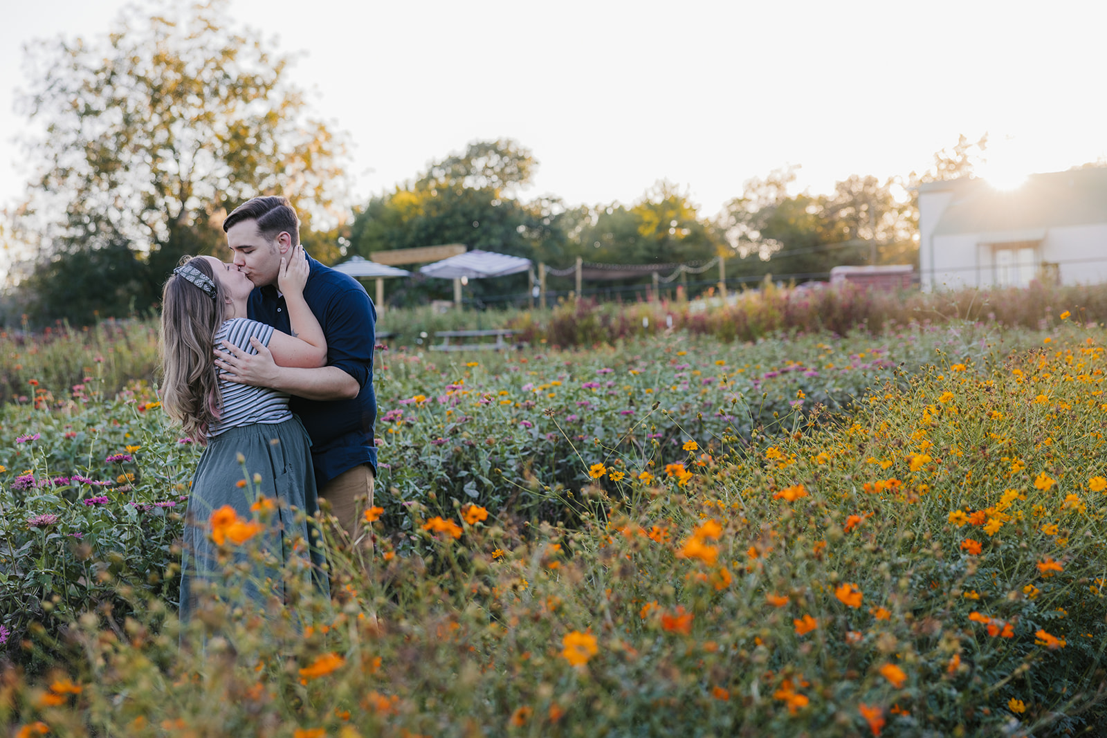 A couple embraces in a field of wildflowers during sunset, with trees and a structure visible in the background.