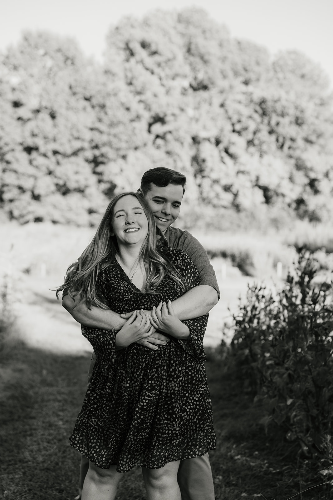 A man and woman stand in a garden, with the man hugging the woman from behind. The woman smiles gently, and there are colorful flowers in the background.