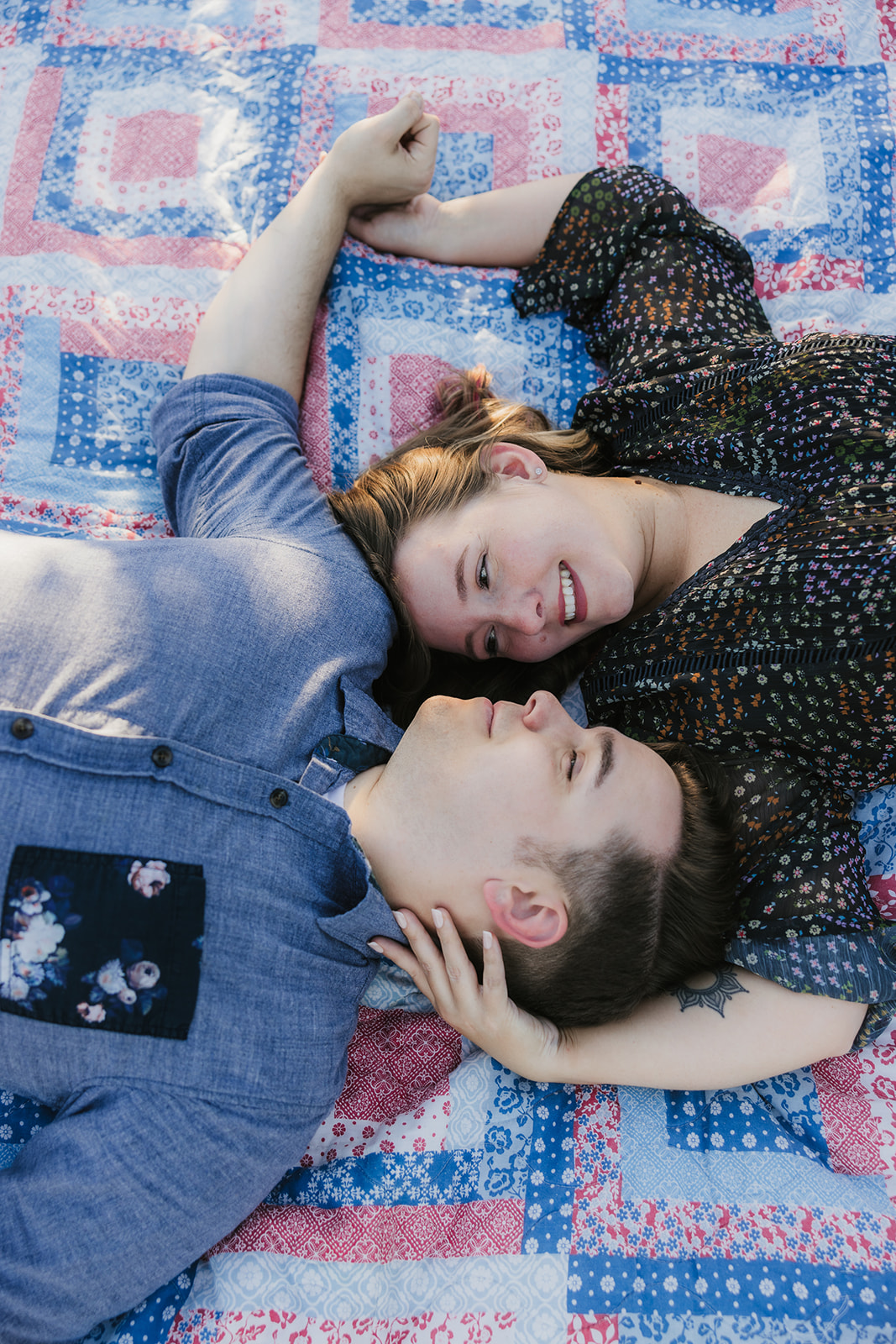 A couple sits on a blanket in a garden, surrounded by plants and trees, enjoying a sunny day.